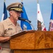 A U.S. Navy officer in Service Khaki uniform is speaking at a podium during an official ceremony. The wooden podium is engraved with 'TACTICAL TRAINING GROUP PACIFIC,' indicating this is a formal military event at that command. Multiple state and organizational flags are displayed in the background.