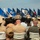 C3F conducts Chief pinning ceremony A formal U.S. Navy military ceremony is taking place outdoors, with uniformed personnel and civilian guests seated in white chairs facing a stage or podium area. Two Navy sailors in tan/khaki service uniforms stand at the front, with a large display of U.S. state flags arranged behind them. The setting appears to be a graduation, change of command, or similar official military ceremony.
