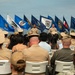 A formal U.S. Navy military ceremony is taking place outdoors, with uniformed personnel and civilian guests seated in white chairs facing a stage or podium area. Two Navy sailors in tan/khaki service uniforms stand at the front, with a large display of U.S. state flags arranged behind them. The setting appears to be a graduation, change of command, or similar official military ceremony.