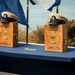 Two wooden shadow boxes or retirement/promotion keepsake boxes are displayed on a navy blue draped table at what appears to be a U.S. Navy ceremony. Each box bears a USN anchor emblem, military qualification badges, an engraved name, and the date 'September 16, 2026', with a white-topped Navy chief petty officer cover (hat) resting on top of each. The setting includes naval flags in the background and appears to be an outdoor waterfront location.