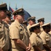 A group of United States Navy Chief Petty Officers are standing in formation during what appears to be a formal military ceremony. They are wearing the standard khaki Service Dress Khaki uniforms with combination covers (peaked caps) bearing gold anchors. Multiple flags are visible in the background, suggesting an official naval event or ceremony.