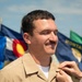 A man in a tan/khaki military uniform is having a pin or insignia affixed to his collar by another person's hand. He is smiling with eyes closed, suggesting a ceremonial or celebratory moment such as a promotion or award ceremony. Multiple U.S. state flags are visible in the background, including Colorado and Montana.