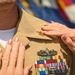 A close-up image showing two sets of hands pinning insignia onto a U.S. Navy officer's khaki uniform during what appears to be a promotion or award ceremony. The uniform displays a Surface Warfare Officer (SWO) badge and multiple service ribbons. The ceremonial pinning suggests a formal military milestone event.