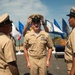 A U.S. Navy chief petty officer pinning/hat ceremony is taking place outdoors, likely at a coastal military installation. The central figure is placing a chief petty officer cover (cap) on his own head while flanked by two senior enlisted personnel in khaki service uniforms. Multiple national and military flags are displayed in the background near what appears to be a ceremonial table.