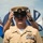 C3F conducts Chief pinning ceremony A U.S. Navy Chief Petty Officer in Service Khaki uniform is adjusting his combination cover (cap) with both hands during what appears to be a formal military ceremony. Multiple flags are visible in the background, including what appears to be state and unit flags. The subject displays multiple service ribbons and a warfare qualification badge on his uniform.