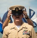 A U.S. Navy Chief Petty Officer in Service Khaki uniform is adjusting his combination cover (cap) with both hands during what appears to be a formal military ceremony. Multiple flags are visible in the background, including what appears to be state and unit flags. The subject displays multiple service ribbons and a warfare qualification badge on his uniform.