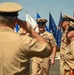 A formal U.S. Navy military ceremony is taking place outdoors, likely a change of command or retirement ceremony. Multiple naval personnel in tan/khaki Service Uniform B are exchanging salutes, with one central figure walking forward while rendering a salute. A color guard with multiple flags, including blue and white naval flags, is visible in the background.