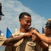 A U.S. Navy promotion or pinning ceremony is taking place outdoors under a blue sky. A smiling sailor in khaki uniform is having insignia or rank devices pinned onto his uniform by three individuals, including fellow sailors and what appears to be a civilian woman. Multiple naval flags are visible in the background.