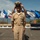 C3F conducts Chief pinning ceremony A U.S. Navy Chief Petty Officer in khaki service uniform is adjusting his combination cover (cap) during what appears to be a promotion or pinning ceremony. He is smiling broadly and standing in front of a row of U.S. state flags, with a table bearing wooden plaques visible to his right. The outdoor setting appears to be a coastal location, likely in California based on the visible California Republic flag.