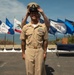 A U.S. Navy Chief Petty Officer in khaki service uniform is adjusting his combination cover (cap) during what appears to be a promotion or pinning ceremony. He is smiling broadly and standing in front of a row of U.S. state flags, with a table bearing wooden plaques visible to his right. The outdoor setting appears to be a coastal location, likely in California based on the visible California Republic flag.