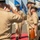 C3F conducts Chief pinning ceremony A formal U.S. Navy military ceremony is taking place outdoors, likely a change of command or retirement ceremony. Personnel in tan/khaki Service Uniform B are exchanging salutes as an officer walks along a red carpet between two rows of sailors. Multiple state and organizational flags are displayed in the background near what appears to be a waterfront location.