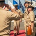 A formal U.S. Navy military ceremony is taking place outdoors, likely a change of command or retirement ceremony. Personnel in tan/khaki Service Uniform B are exchanging salutes as an officer walks along a red carpet between two rows of sailors. Multiple state and organizational flags are displayed in the background near what appears to be a waterfront location.