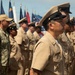 A formation of U.S. Navy personnel standing at attention during a formal military ceremony. Personnel are wearing a mix of Service Khaki uniforms with combination covers and Navy Working Uniform (NWU) camouflage pattern. Multiple military and state flags are visible in the background.