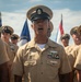 A group of U.S. Navy personnel in Service Khaki uniforms are assembled outdoors, likely during a formal ceremony or parade. The central figure, a senior enlisted sailor wearing a combination cover with an anchor insignia, displays multiple service ribbons and warfare qualification badges on his uniform. Several flags, including what appears to be a Commonwealth flag, are visible in the background.