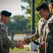 Two U.S. Army soldiers in MultiCam (OCP) uniforms are engaged in a handshake during what appears to be a formal military ceremony or award presentation. The soldier on the right, wearing a black beret with a general officer star device, appears to be presenting or receiving something while shaking hands. Additional uniformed personnel are partially visible on both sides of the frame.