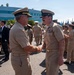 Two U.S. Navy senior enlisted personnel are shaking hands in the foreground during what appears to be a formal military ceremony or change of command event. Both are wearing khaki service uniforms with combination covers, and the individual on the right displays multiple service ribbons. A formation of Navy personnel in matching uniforms stands in the background alongside civilian spectators.