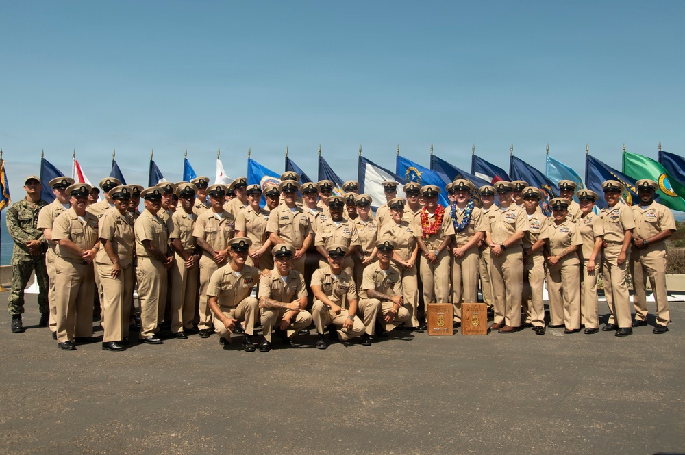 A large group of U.S. Navy personnel in tan/khaki service uniforms pose for a formal group photograph outdoors. They are arranged in multiple rows with a display of numerous military flags and pennants behind them. One individual near the center is wearing a colorful lei, suggesting a ceremonial or special occasion, possibly in Hawaii.