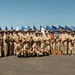 A large group of U.S. Navy personnel in tan/khaki service uniforms pose for a formal group photograph outdoors. They are arranged in multiple rows with a display of numerous military flags and pennants behind them. One individual near the center is wearing a colorful lei, suggesting a ceremonial or special occasion, possibly in Hawaii.