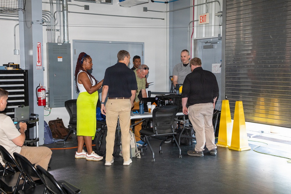 A group of approximately seven people are gathered in what appears to be an industrial or workshop facility, clustered around a table with equipment. The setting features exposed ductwork, a fire extinguisher, electrical panel, and yellow safety cones near a large roll-up door. The gathering has the appearance of a briefing, demonstration, or training session.