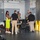 xTechAutonomy Phase II Live Demonstrations A group of approximately seven people are gathered in what appears to be an industrial or workshop facility, clustered around a table with equipment. The setting features exposed ductwork, a fire extinguisher, electrical panel, and yellow safety cones near a large roll-up door. The gathering has the appearance of a briefing, demonstration, or training session.
