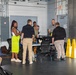 A group of approximately seven people are gathered in what appears to be an industrial or workshop facility, clustered around a table with equipment. The setting features exposed ductwork, a fire extinguisher, electrical panel, and yellow safety cones near a large roll-up door. The gathering has the appearance of a briefing, demonstration, or training session.