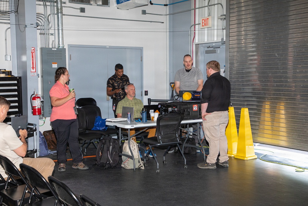 A group of approximately six people are gathered in what appears to be an industrial or garage-style workspace, likely during a meeting or training session. Several individuals are clustered around a folding table with a laptop and various equipment, while others stand or sit nearby. The setting includes safety equipment, tool storage, and large roll-up doors typical of a maintenance or operations facility.