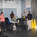 A group of approximately six people are gathered in what appears to be an industrial or garage-style workspace, likely during a meeting or training session. Several individuals are clustered around a folding table with a laptop and various equipment, while others stand or sit nearby. The setting includes safety equipment, tool storage, and large roll-up doors typical of a maintenance or operations facility.