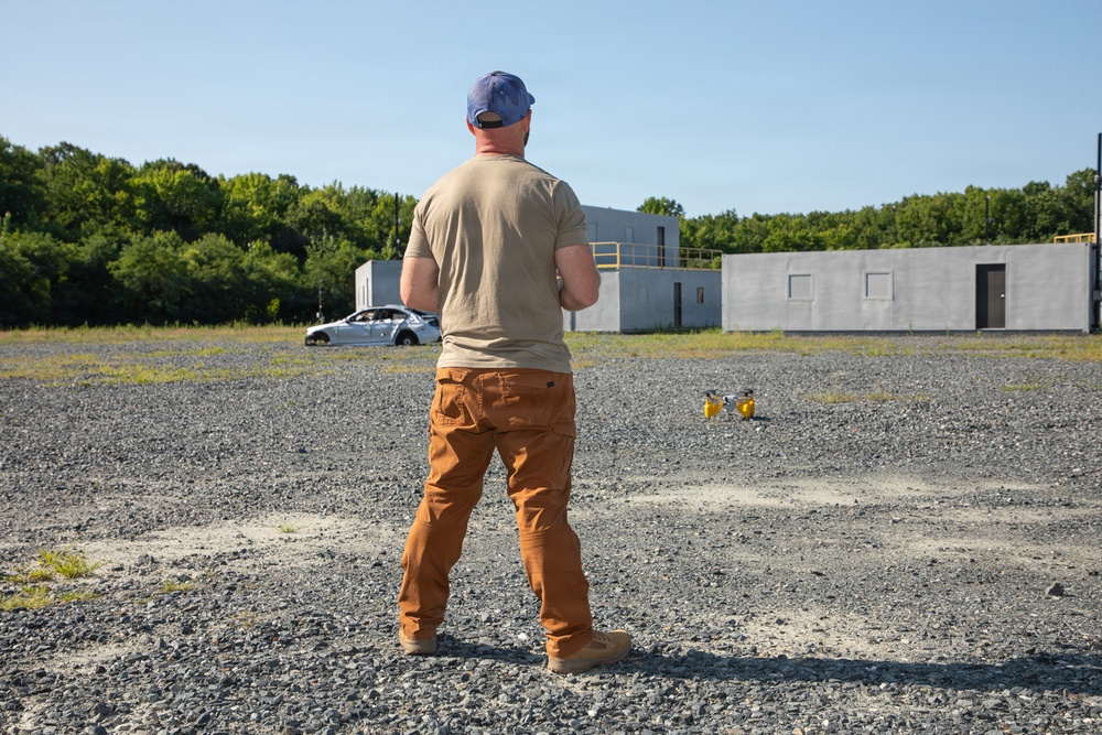 A man wearing a backwards blue cap, tan t-shirt, and brown cargo pants stands on a gravel surface facing away from the camera. He appears to be observing or operating a small drone on the ground in front of him. The background includes concrete training structures, a damaged vehicle, and a tree line under clear skies.