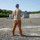 xTechAutonomy Phase II Live Demonstrations A man wearing a backwards blue cap, tan t-shirt, and brown cargo pants stands on a gravel surface facing away from the camera. He appears to be observing or operating a small drone on the ground in front of him. The background includes concrete training structures, a damaged vehicle, and a tree line under clear skies.