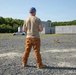 A man wearing a backwards blue cap, tan t-shirt, and brown cargo pants stands on a gravel surface facing away from the camera. He appears to be observing or operating a small drone on the ground in front of him. The background includes concrete training structures, a damaged vehicle, and a tree line under clear skies.
