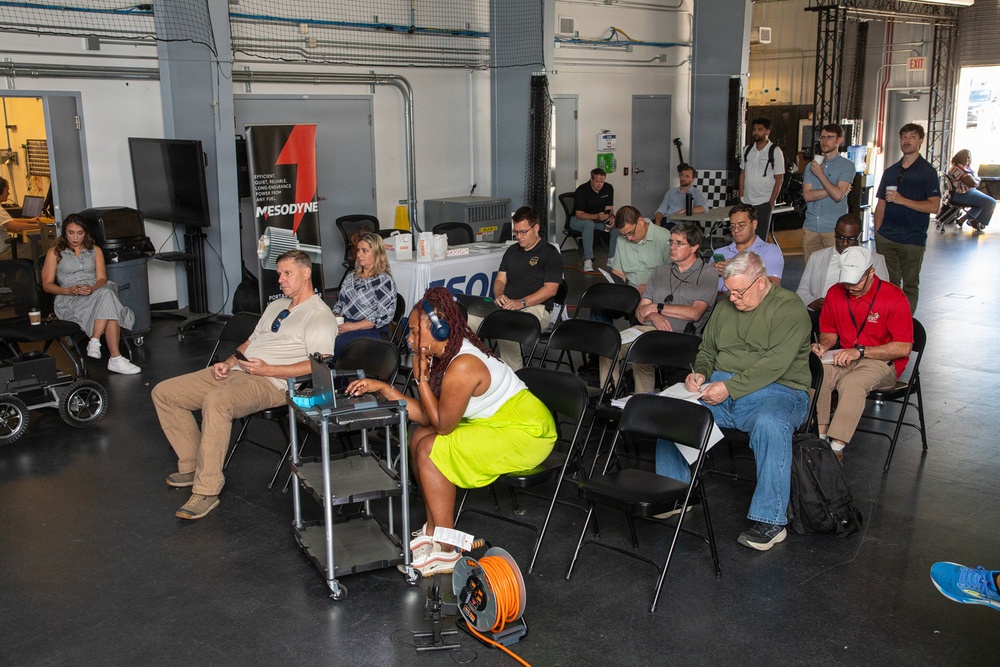A group of approximately 15-20 people are gathered in what appears to be an industrial or garage-style venue for a presentation or informational event. A banner for 'Mesodyne' is prominently displayed at the front, suggesting this is a company demonstration or pitch event. Attendees are seated in folding chairs while some stand along the periphery, with several taking notes.
