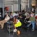 A group of approximately 15-20 people are gathered in what appears to be an industrial or garage-style venue for a presentation or informational event. A banner for 'Mesodyne' is prominently displayed at the front, suggesting this is a company demonstration or pitch event. Attendees are seated in folding chairs while some stand along the periphery, with several taking notes.