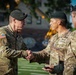Two U.S. Army soldiers in OCP (Operational Camouflage Pattern) uniforms are shaking hands in what appears to be a formal military ceremony or recognition event. A third soldier in the same uniform is visible in the right foreground. The setting appears to be an outdoor athletic field or stadium.