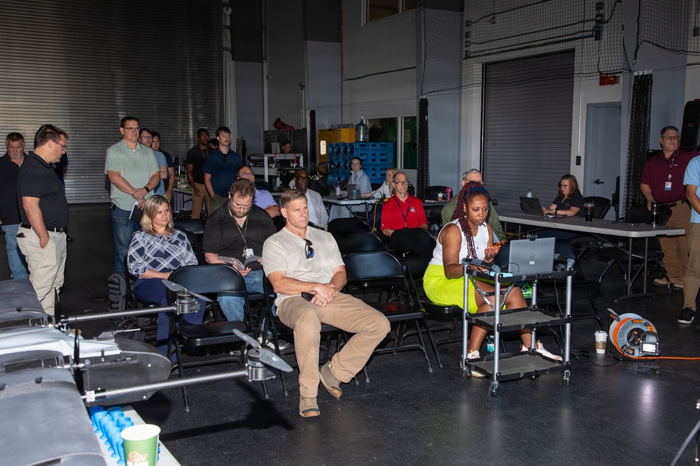 A group of approximately 20-25 people are gathered in what appears to be an industrial or hangar-style facility for a presentation or briefing. Attendees are seated in folding chairs arranged in rows, while some individuals stand along the perimeter. A drone or UAV is visible in the foreground, suggesting the event is related to unmanned aerial systems or aerospace technology.