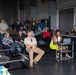 A group of approximately 20-25 people are gathered in what appears to be an industrial or hangar-style facility for a presentation or briefing. Attendees are seated in folding chairs arranged in rows, while some individuals stand along the perimeter. A drone or UAV is visible in the foreground, suggesting the event is related to unmanned aerial systems or aerospace technology.