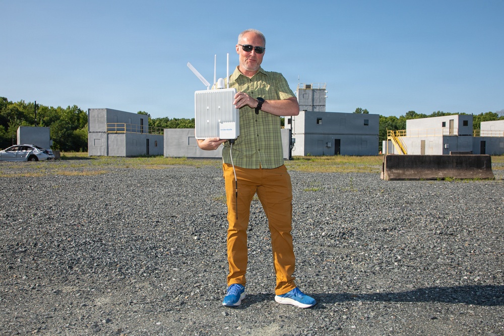 A middle-aged man stands on a gravel surface holding a white wireless networking device with multiple antennas. He is positioned in front of what appears to be a training or testing facility with concrete block structures. The setting suggests a field demonstration or equipment testing scenario.