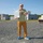 xTechAutonomy Phase II Live Demonstrations A middle-aged man stands on a gravel surface holding a white wireless networking device with multiple antennas. He is positioned in front of what appears to be a training or testing facility with concrete block structures. The setting suggests a field demonstration or equipment testing scenario.