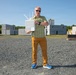 A middle-aged man stands on a gravel surface holding a white wireless networking device with multiple antennas. He is positioned in front of what appears to be a training or testing facility with concrete block structures. The setting suggests a field demonstration or equipment testing scenario.