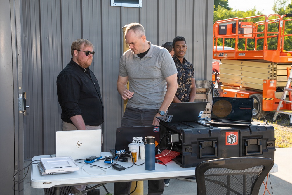 Three men are gathered around an outdoor workstation table set up against a metal building. The table holds multiple laptops, a rugged equipment case, beverages, and various accessories, suggesting a field operations or technical demonstration setup. A JLG scissor lift is visible in the background to the right.