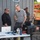 xTechAutonomy Phase II Live Demonstrations Three men are gathered around an outdoor workstation table set up against a metal building. The table holds multiple laptops, a rugged equipment case, beverages, and various accessories, suggesting a field operations or technical demonstration setup. A JLG scissor lift is visible in the background to the right.