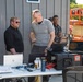 Three men are gathered around an outdoor workstation table set up against a metal building. The table holds multiple laptops, a rugged equipment case, beverages, and various accessories, suggesting a field operations or technical demonstration setup. A JLG scissor lift is visible in the background to the right.
