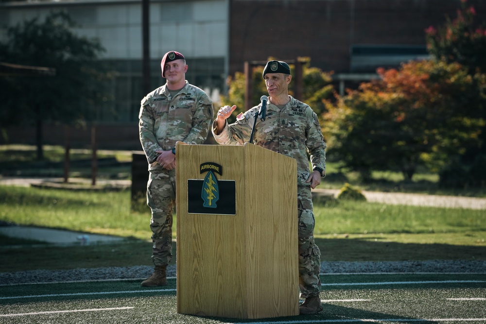 Two U.S. Army soldiers in MultiCam (OCP) uniforms are positioned at a wooden podium bearing the U.S. Army Special Forces (Airborne) insignia. One soldier is speaking at the microphone while the other stands at ease in the background. The setting appears to be an outdoor ceremony on a athletic field at a military installation.