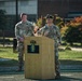 Two U.S. Army soldiers in MultiCam (OCP) uniforms are positioned at a wooden podium bearing the U.S. Army Special Forces (Airborne) insignia. One soldier is speaking at the microphone while the other stands at ease in the background. The setting appears to be an outdoor ceremony on a athletic field at a military installation.