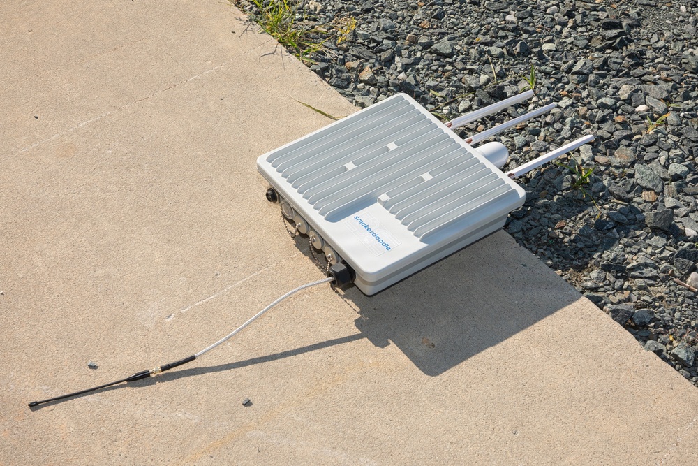 The image shows a white outdoor networking or wireless communication device branded 'Snickerdoodle' resting on a concrete surface near a gravel border. The device has multiple antennas extending from one side and a white cable connected to it with an additional antenna lying on the concrete. The equipment appears to be an outdoor-rated enclosure, likely a wireless access point or IoT gateway.