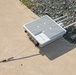 The image shows a white outdoor networking or wireless communication device branded 'Snickerdoodle' resting on a concrete surface near a gravel border. The device has multiple antennas extending from one side and a white cable connected to it with an additional antenna lying on the concrete. The equipment appears to be an outdoor-rated enclosure, likely a wireless access point or IoT gateway.