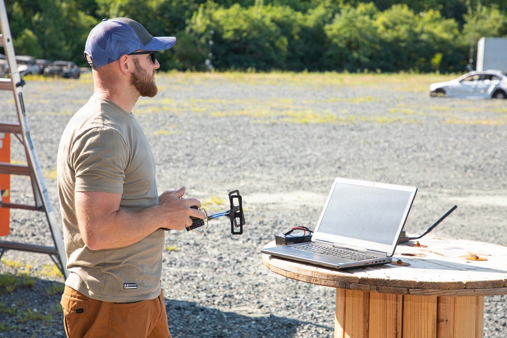 A bearded man wearing a blue cap, sunglasses, and a tan t-shirt stands outdoors on a gravel surface holding what appears to be a drone remote controller. He is positioned next to a makeshift wooden cable spool table on which a laptop computer and some electronic components are placed. The setting appears to be an open field or testing area with trees and a vehicle visible in the background.