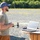 xTechAutonomy Phase II Live Demonstrations A bearded man wearing a blue cap, sunglasses, and a tan t-shirt stands outdoors on a gravel surface holding what appears to be a drone remote controller. He is positioned next to a makeshift wooden cable spool table on which a laptop computer and some electronic components are placed. The setting appears to be an open field or testing area with trees and a vehicle visible in the background.