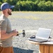 A bearded man wearing a blue cap, sunglasses, and a tan t-shirt stands outdoors on a gravel surface holding what appears to be a drone remote controller. He is positioned next to a makeshift wooden cable spool table on which a laptop computer and some electronic components are placed. The setting appears to be an open field or testing area with trees and a vehicle visible in the background.