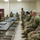 A group of U.S. Army soldiers in OCP (Operational Camouflage Pattern) uniforms are gathered in a classroom or briefing room setting. One soldier appears to be standing and addressing the group, suggesting a formal briefing, training session, or after-action review. The room contains tables with laptops, a whiteboard, and a projector.