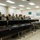 A military classroom setting where a senior soldier or officer is addressing a group of approximately 20 uniformed personnel seated at tiered desks. All individuals are wearing US Army Operational Camouflage Pattern (OCP) uniforms. The instructor appears to be conducting a briefing or training session in what appears to be a dedicated military training facility.
